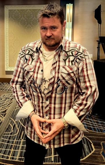 Man in a patterned shirt stands confidently with hands clasped in a stylish room.