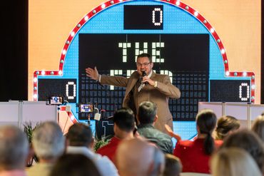 A lively host engages an audience in a game show setting with a colorful scoreboard behind.