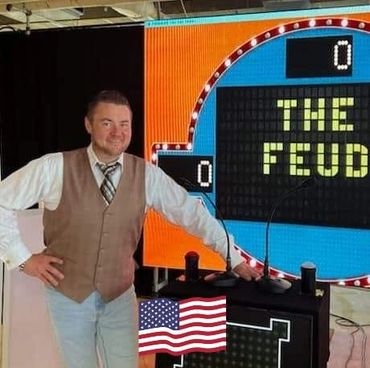 Man standing beside a game show podium with 'The Feud' on the screen.