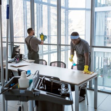 Two janitors cleaning an office with large windows and desks.