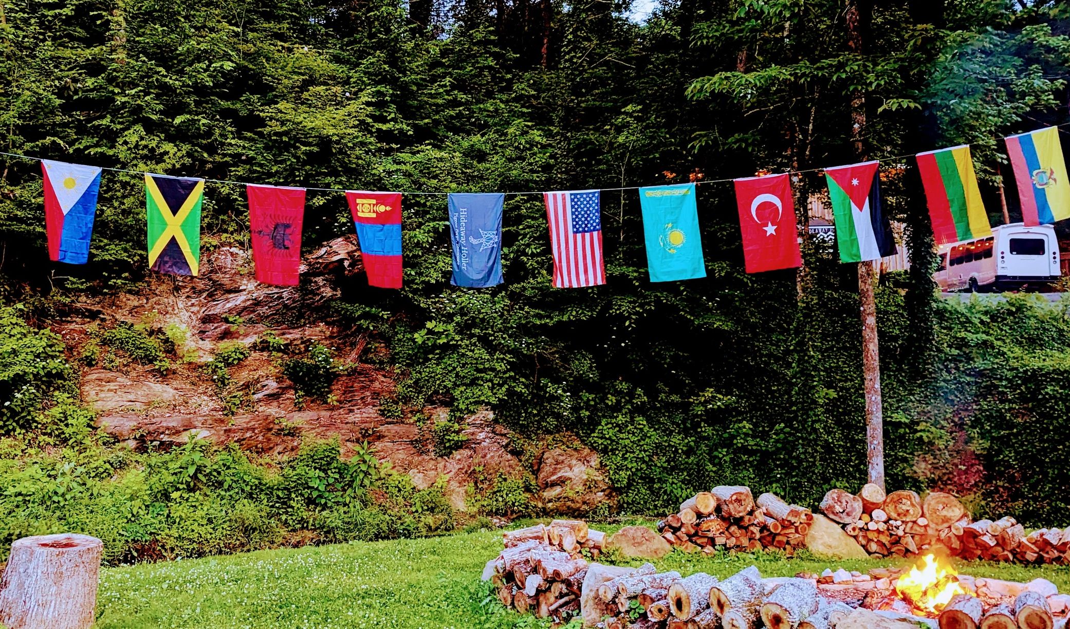 A row of international flags hanging outdoors with a campfire and forest background.