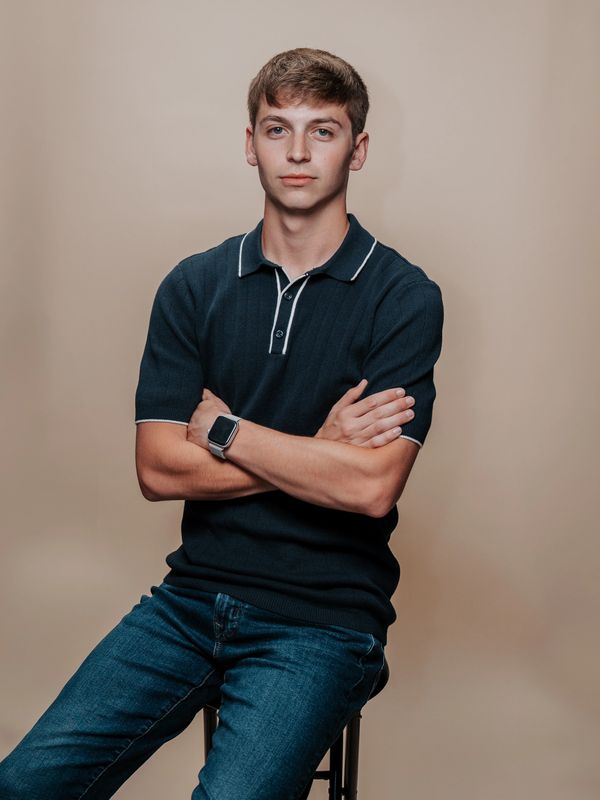 Young man sitting on a stool with arms crossed, wearing a black polo and jeans.
