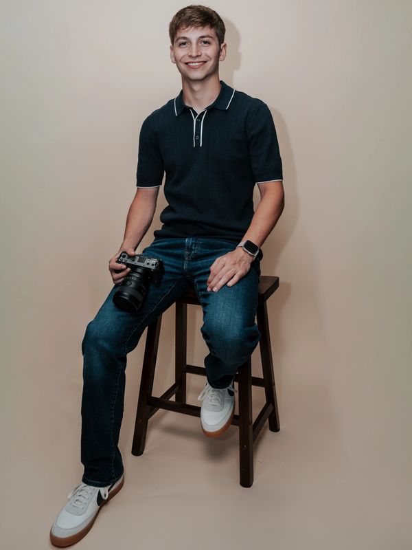 Young man sitting on a stool holding a camera, smiling.