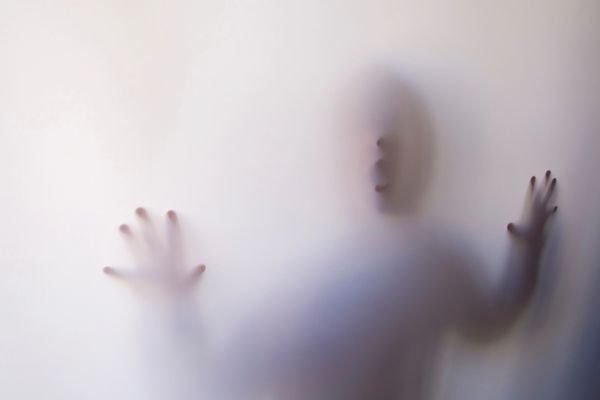 A ghosted image of a human person looking through shaded glass & pressing their hands onto a window.