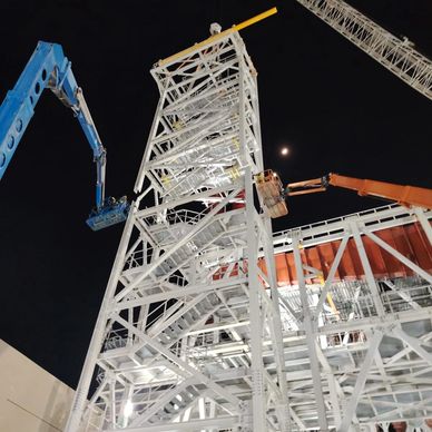 Construction workers on lifts working on a tall white metal structure at night.