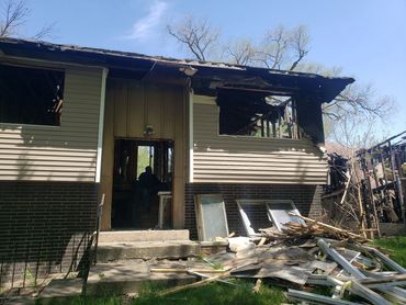Burned house exterior with debris scattered outside under clear sky.