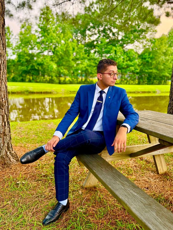 Man in a blue suit sitting on a picnic bench by a pond.