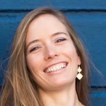 Smiling woman with long hair and geometric earrings against a blue wall.