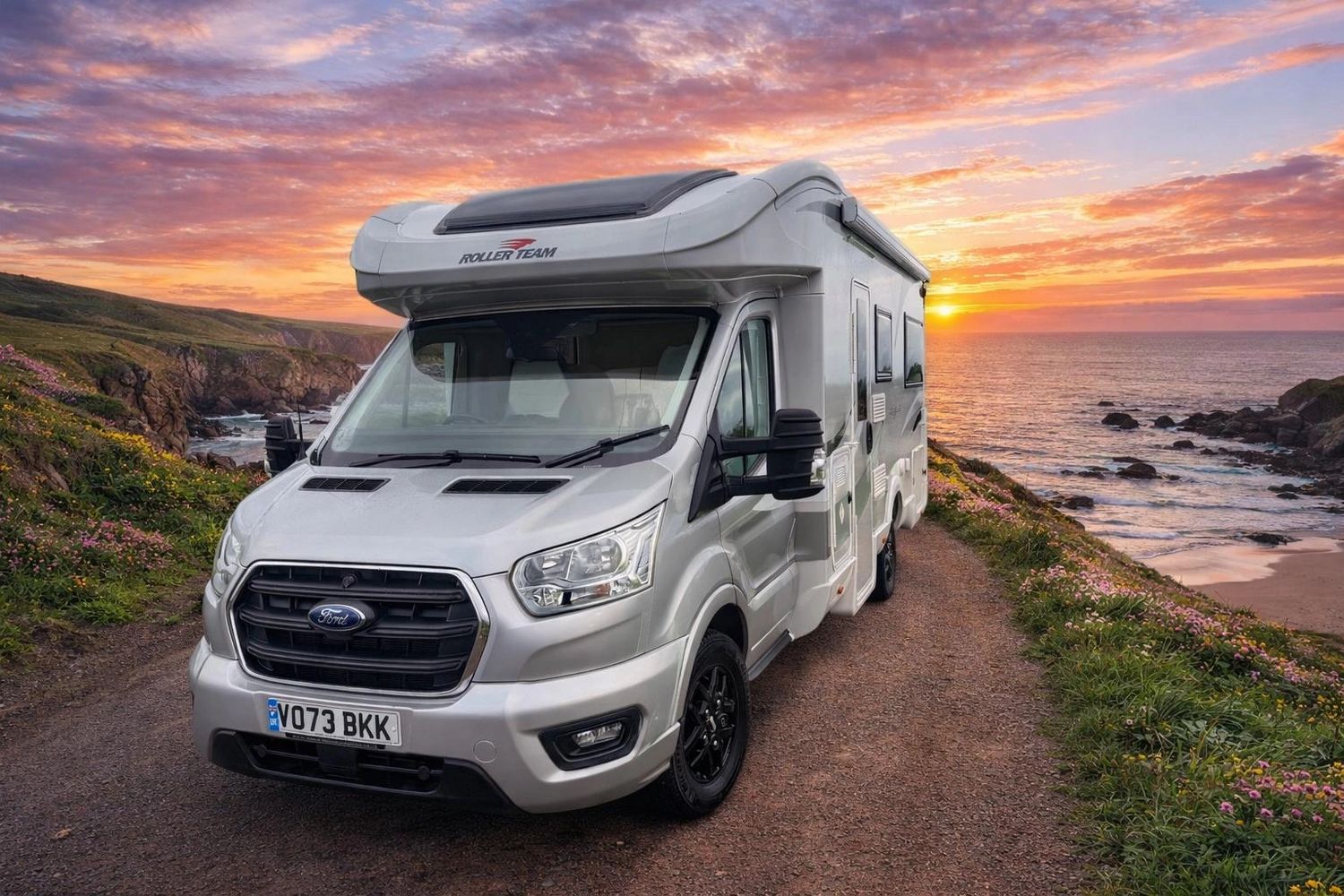 Silver motorhome parked on a coastal road at sunset.