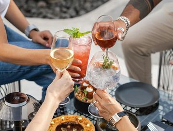 Four people clinking glasses with cocktails and wine over a table with food.