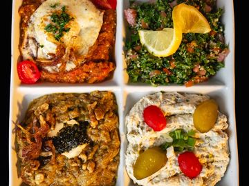 A divided plate with four different Middle Eastern dishes, garnished with eggs, lemon, pickles, and herbs.