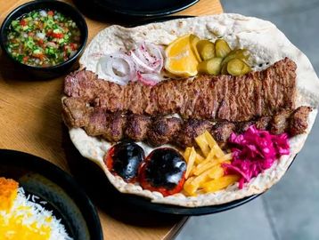 Grilled meat kebabs served with flatbread, pickles, onions, lemon, grilled tomatoes, fries, and purple cabbage.