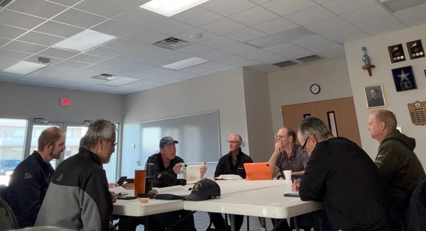 A group of seven men engaged in a meeting around a white table in a conference room.