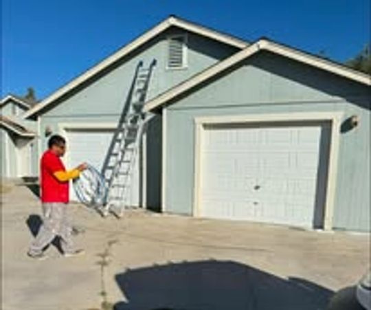 Man in red shirt walking past a light blue house with a ladder and two white garage doors