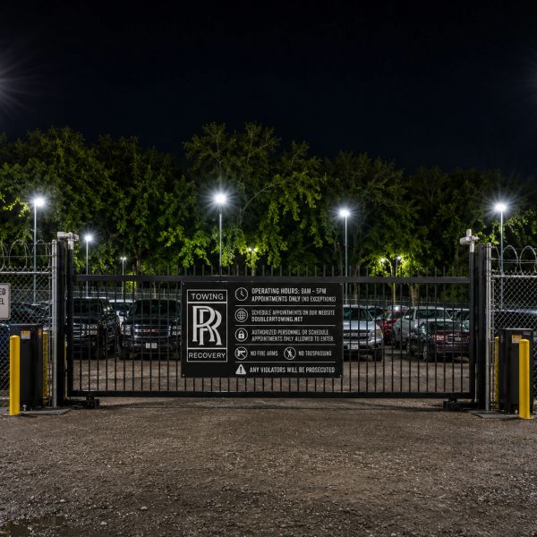 Gated towing recovery lot at night with illuminated sign and parked cars.