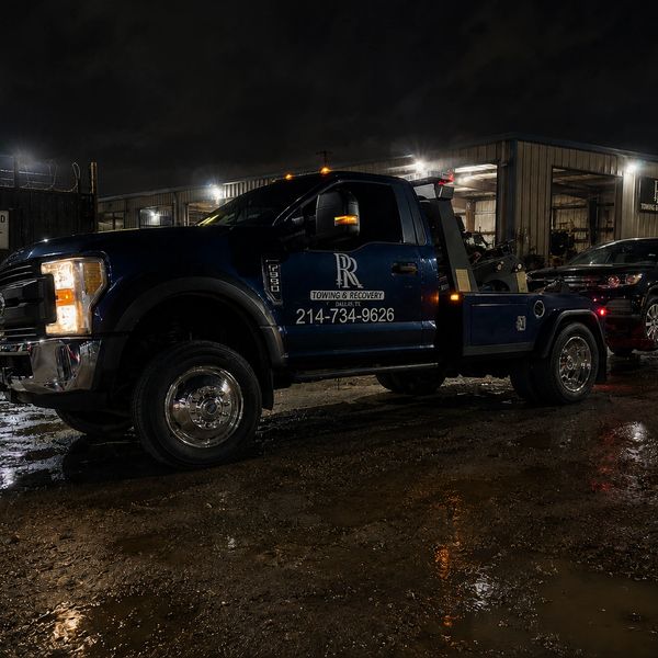 A dark blue tow truck parked outside a lit garage at night.