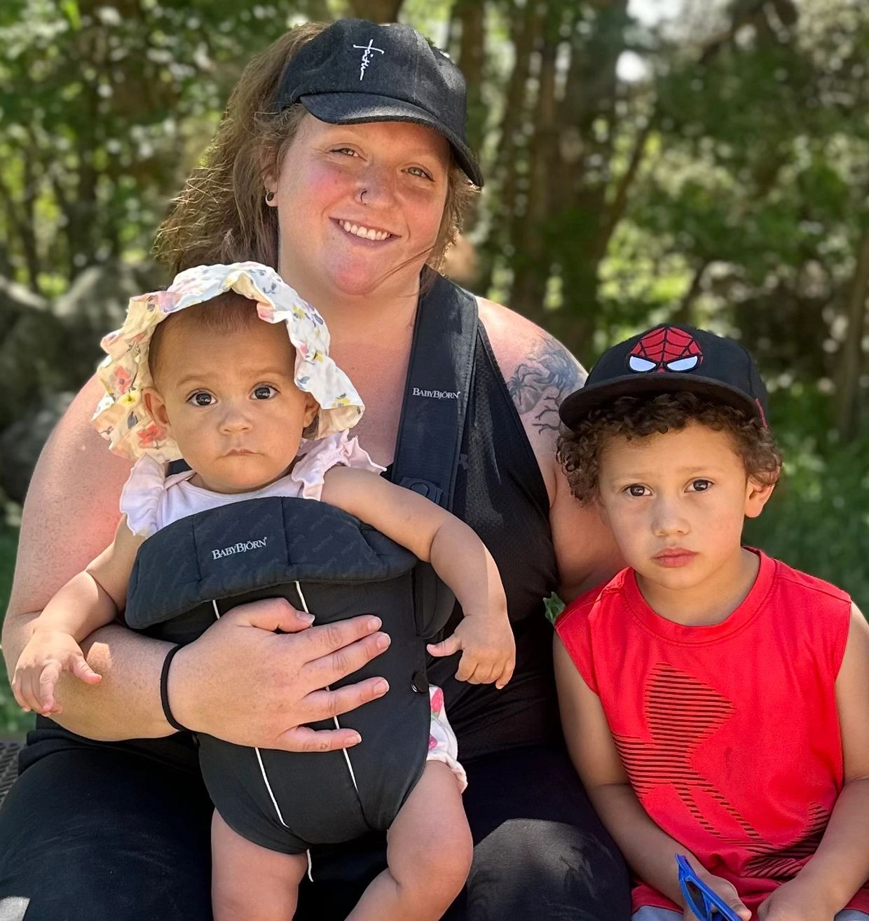 A woman with two children outdoors, one baby in a carrier and an older boy in a red shirt and Spider-Man hat.