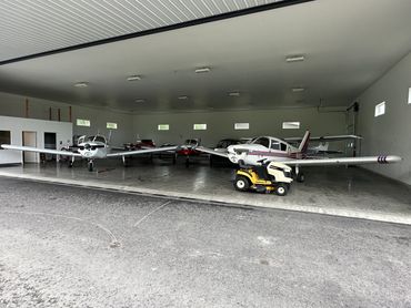 Small airplanes parked inside a spacious hangar with a lawnmower in front.
