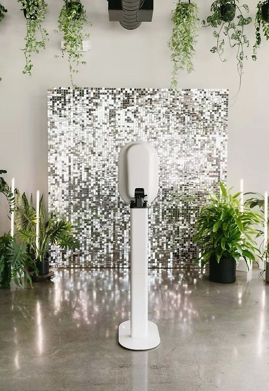 White sanitizer dispenser with silver backdrop and surrounding green plants.