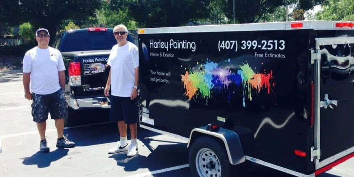 Two men stand beside a black trailer and truck advertising Harley Painting services.