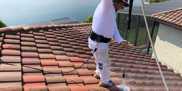Worker pressure washing a tiled roof while secured with safety gear.