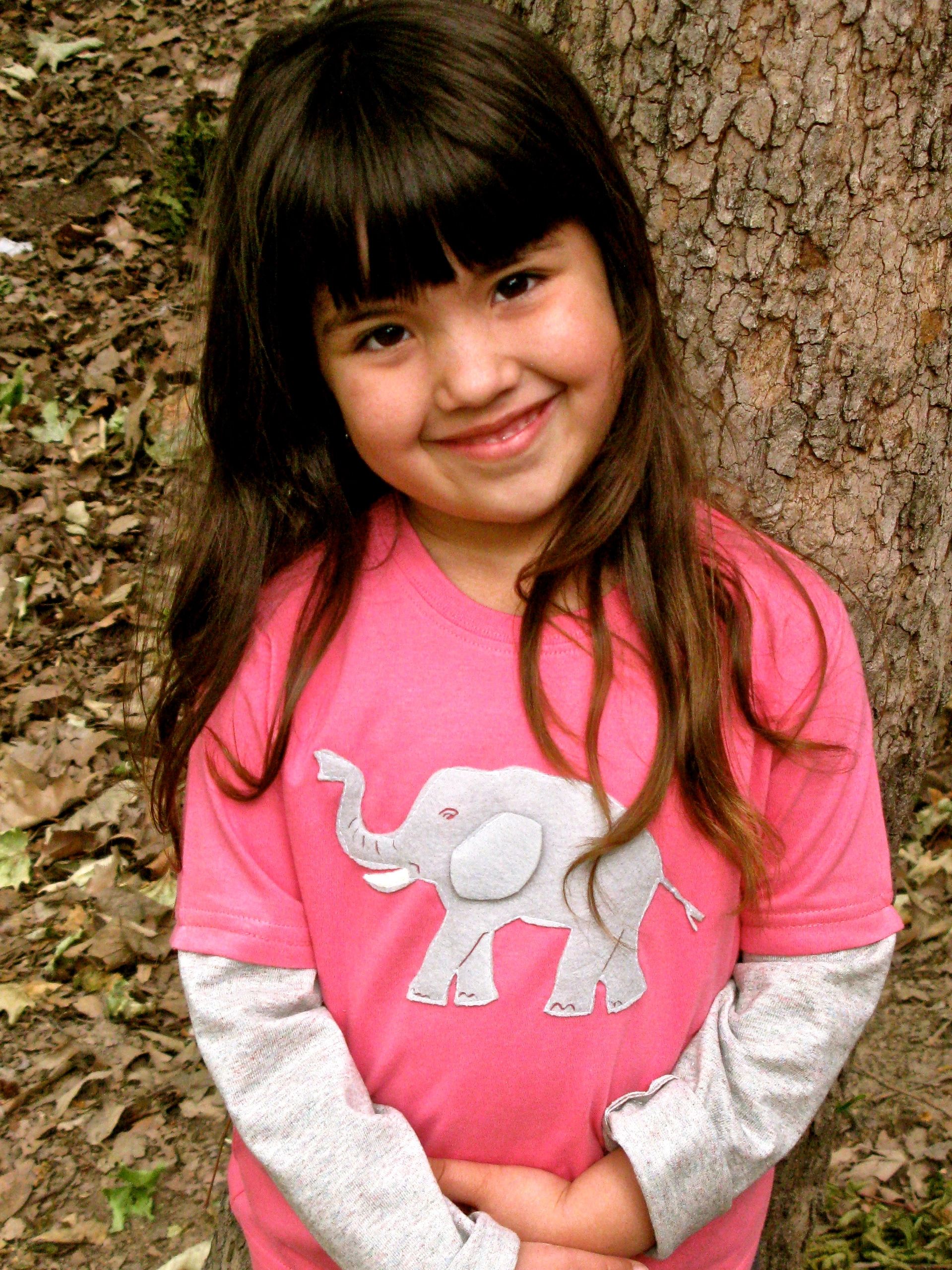 Smiling girl in a pink shirt with an elephant design leaning against a tree.