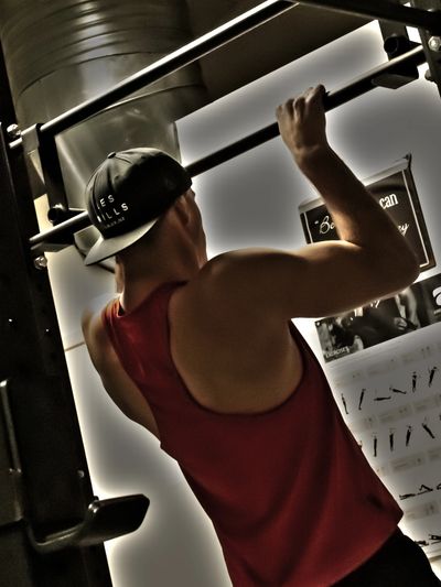 Man doing pull-ups in a gym wearing a red tank top and a backwards cap.