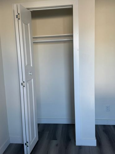 Empty white closet with folding door and dark wood flooring.