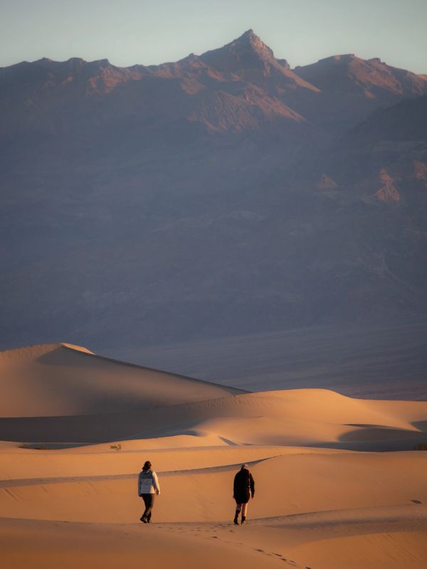 Two people walking on sand dunes with mountains in the background at sunset.