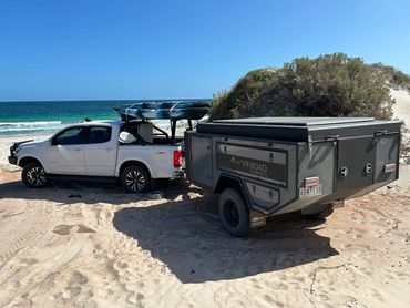 White 4x4 ute towing an off-road camper trailer on a sandy beach with ocean in the background.