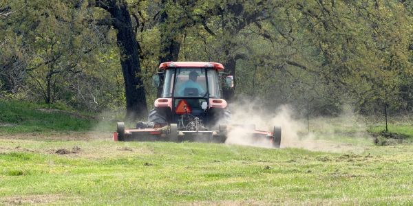 A tractor mowing a dusty field surrounded by trees.