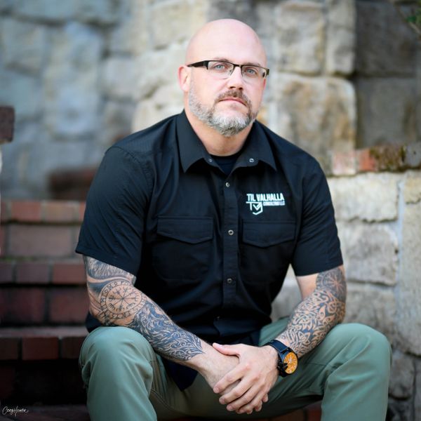 Man with glasses and tattoos sits on steps against stone wall.