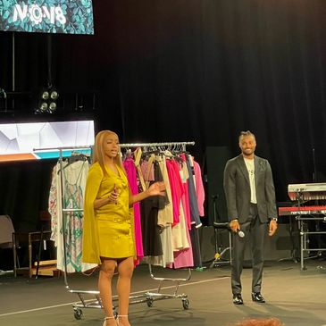 Two presenters on stage with clothing rack and microphone.