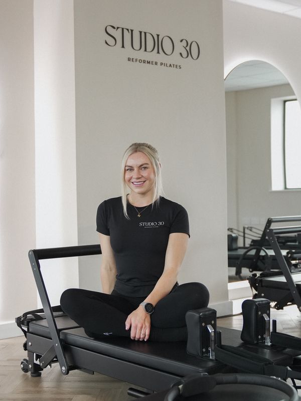 Smiling woman in black Pilates attire sitting on reformer machine at Studio 30.
