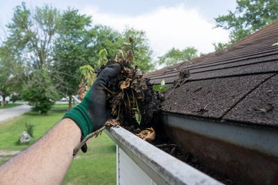 Hand cleaning leaves and debris from a clogged gutter on a roof.