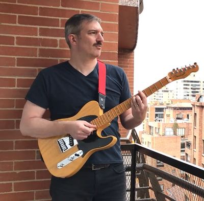 Tall, mustachioed, white man on a balcony playing a natural wood tone Fender electric guitar