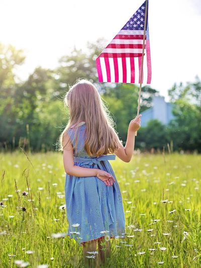 Blonde child in a blue dress holding an American Flag in a field