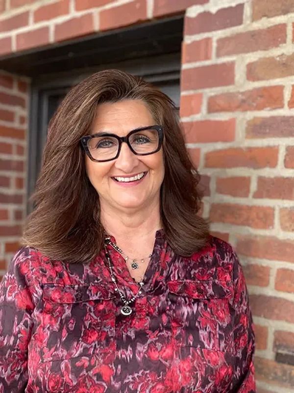 Therapist Sharon Thomsen, with a warm, inviting smile, glasses in a patterned red blouse against a brick wall.