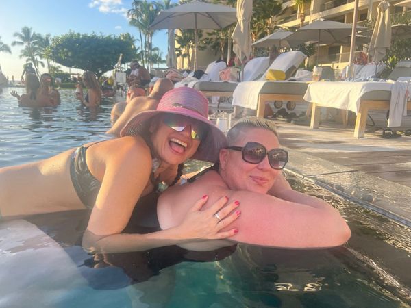Two women relaxing and smiling in a pool at a sunny resort with palm trees and lounge chairs.