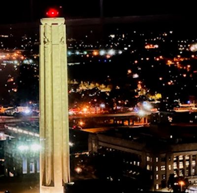 Tall illuminated tower with a red light atop against a city nightscape.