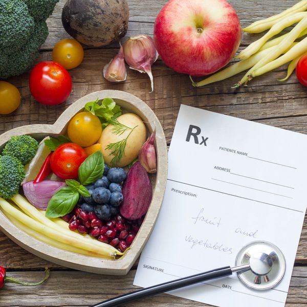 Heart-shaped bowl filled with fresh fruits and vegetables beside a prescription note and stethoscope.