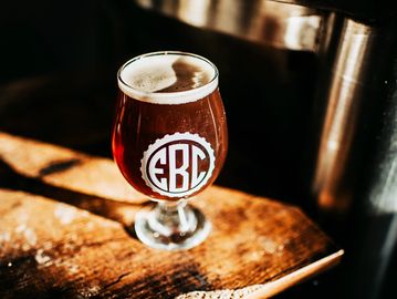 A picture of a dark beer in a tulip glass with kegs behind it.