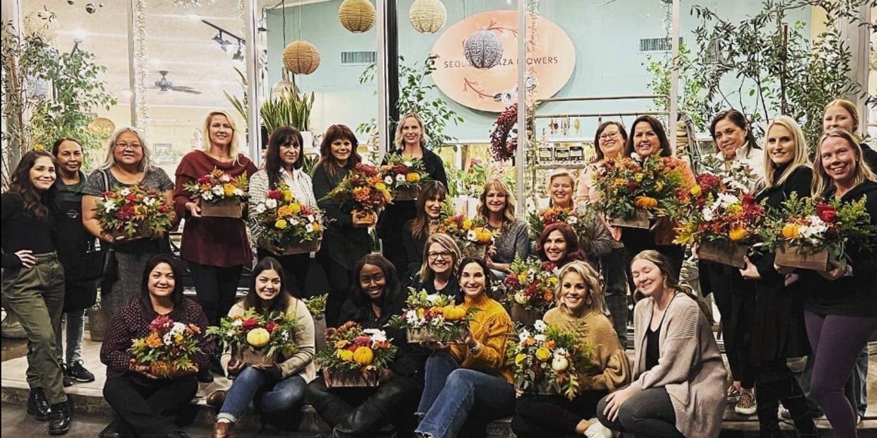Group of smiling women holding flower arrangements indoors.