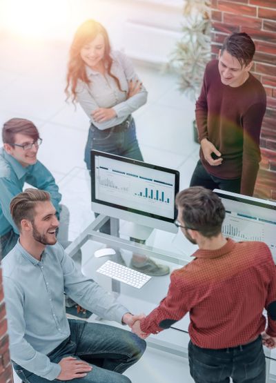 Colleagues shaking hands in a bright office with data charts on screens.