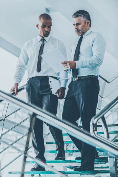 Two businessmen reviewing documents while descending stairs in a modern office.