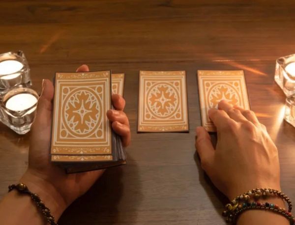 Hands holding and selecting tarot cards on a wooden table lit by candles.