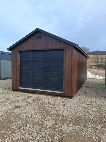 Brown wooden garage with a black garage door on gravel ground.