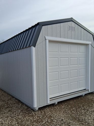 Gray storage shed with a white garage door on a gravel ground.
