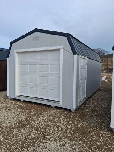 White storage shed with a black roof and a garage door on gravel.
