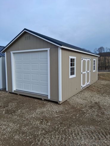 Tan storage shed with white trim, garage door, and windows on gravel ground.